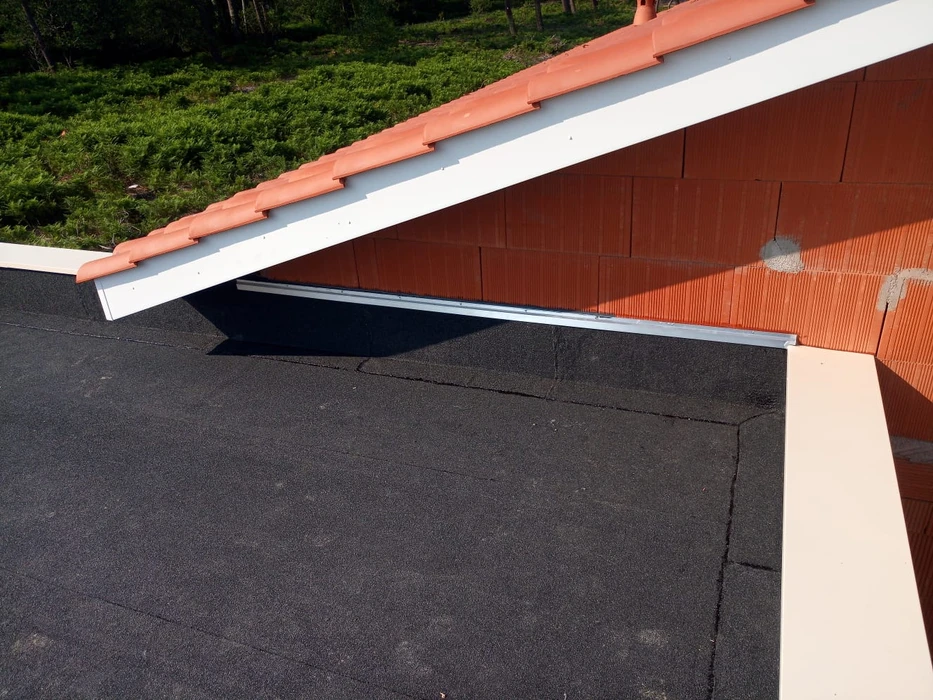 Jonction entre toiture en pente couverte de tuiles en terre cuite et toiture-terrasse plate avec membrane bitumineuse granulée noire