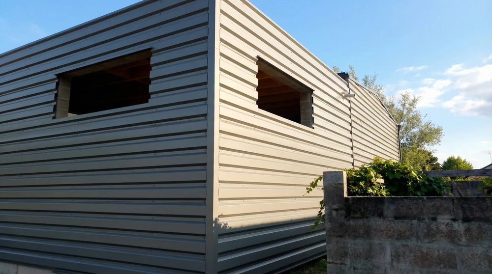 Bâtiment en cours de construction, vue extérieure angle, bardage métallique horizontal gris clair type acier prélaqué, deux ouvertures de fenêtres brutes sans menuiseries avec encadrements bois apparents